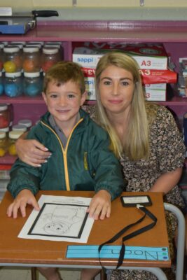A smiling child in a green jacket sits at a school desk with a woman in a floral dress. Art supplies and colorful shelves are in the background. Warm, supportive atmosphere.