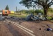 Charred vehicle scattered across a sunny road after a crash, surrounded by debris. Emergency vehicles and personnel are attending the scene.