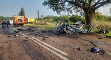 Charred vehicle scattered across a sunny road after a crash, surrounded by debris. Emergency vehicles and personnel are attending the scene.