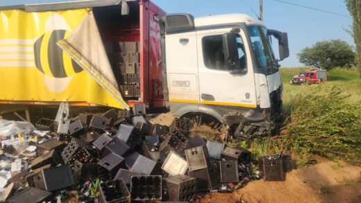 A truck with a yellow and red trailer has collided with vegetation, spilling black crates and bottles onto the ground. In the background, a fire truck is visible.