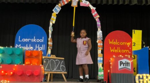 A young girl in a school uniform stands on stage holding a graduation bell under a colorful arch. Background features colorful decorations and "Welcome Welkom" signs, creating a celebratory and festive atmosphere.