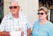 Elderly man with a bandaged hand and sunglasses stands outside with a woman in a blue shirt and sunglasses, both smiling calmly in a sunny setting.