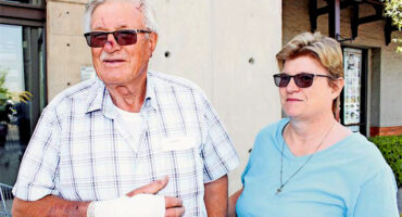 Elderly man with a bandaged hand and sunglasses stands outside with a woman in a blue shirt and sunglasses, both smiling calmly in a sunny setting.
