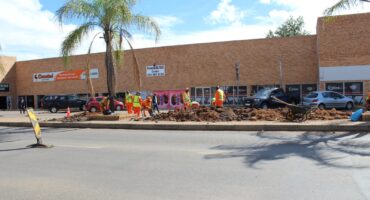Workers in bright vests are digging on a city street median under a palm tree. Shops in the background; roadwork signs and tools scattered around.