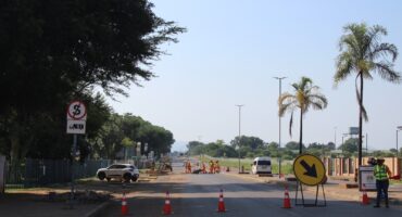 Road construction scene with workers in orange uniforms, traffic cones, and directional signs. A van and parked car are visible under a clear sky.
