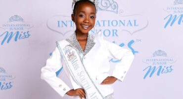 Young girl in a sparkly silver and white outfit with a crown, wearing a "Miss South Africa" sash, smiles confidently against a pageant backdrop.