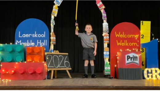 Young boy in school uniform stands on stage, holding a bell under an arch of colorful pencils. Background features large toy blocks, "Laerskool Marble Hall" sign, and "Welcome Welkom" in bold letters. A small chalkboard displays "2026." Vibrant and festive atmosphere.