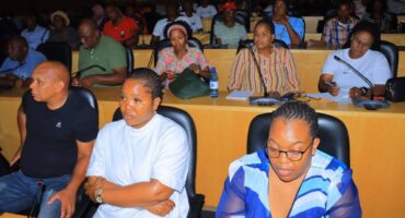 A group of people sit attentively in a conference room with microphones on tables. Some take notes, conveying focus and engagement in the discussion.