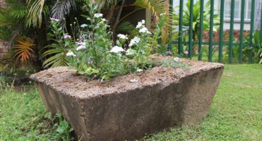 Concrete block with white and purple flowers set in a lush garden. Surrounded by green grass, ferns, and a metal fence in a serene outdoor setting.