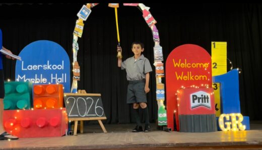 Young boy in school uniform holds a bell under a colorful arch on stage. Bright props include blocks, a "Welcome" sign, and "2026" in chalk. Festive and welcoming atmosphere.