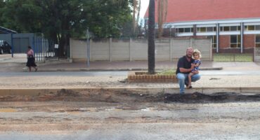 A man crouches on a sidewalk with a child in his arms, smiling near a tree. Behind them, a road with a visible pothole and a red-roofed building.