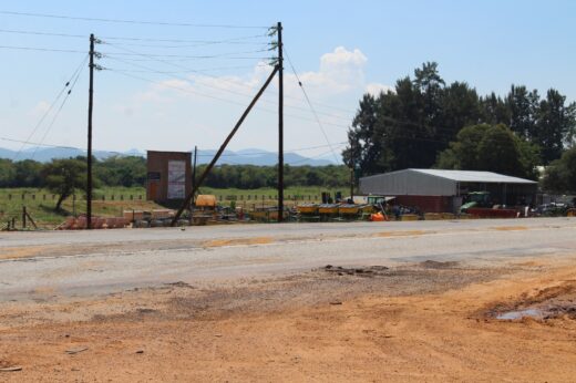 A rural service station with tilted power lines and dusty road in foreground. A metal building, equipment, and trees appear with hills in the distance.