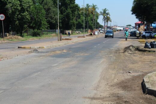 A quiet urban road lined with trees, showing construction signs, vehicles, and a pedestrian. A speed limit sign reads 30 km/h. Sunny day.