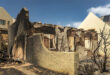 Charred remains of a burnt building with crumbling walls, surrounded by debris and a scorched tree. Adjacent intact houses highlight the devastation.