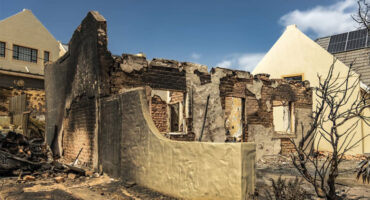 Charred remains of a burnt building with crumbling walls, surrounded by debris and a scorched tree. Adjacent intact houses highlight the devastation.