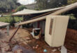 Collapsed patio roof with debris scattered on tiled floor. A fridge is visible next to fallen beams, while a garden and table lie in the background. Mood is chaotic.