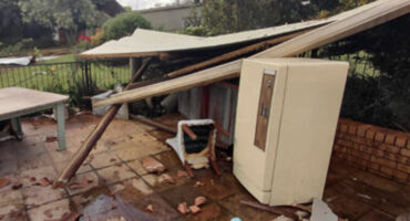 Collapsed patio roof with debris scattered on tiled floor. A fridge is visible next to fallen beams, while a garden and table lie in the background. Mood is chaotic.