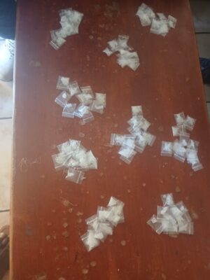 Bags of white powder are scattered on a wooden table with a worn surface. Sneakers and a foot are partially visible at the edges of the image.