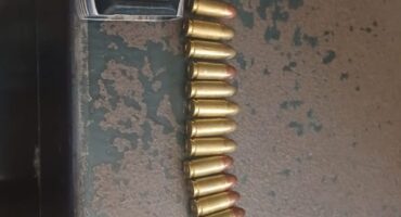 A silver handgun with a black grip lies on a worn metal surface, alongside a row of bullets and an empty magazine clip, evoking themes of tension and danger.