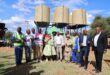 A group of people stand together smiling in front of large elevated water tanks. The scene is outdoors under a blue sky, conveying a sense of achievement.