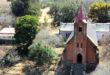 Aerial view of a rustic red brick church with a tall, pointed steeple and cross, surrounded by lush greenery and a small building, evoking tranquility.