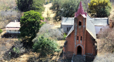 Aerial view of a rustic red brick church with a tall, pointed steeple and cross, surrounded by lush greenery and a small building, evoking tranquility.