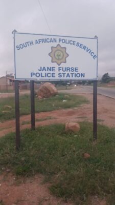 Sign for Jane Furse Police Station in South Africa, standing on grassy ground near a road. Overcast sky suggests a calm, subdued atmosphere.