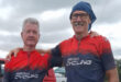 Two elderly men in matching red and blue cycling jerseys stand smiling beside a bike on a grassy field. A red van is parked in the background. Cloudy sky.
