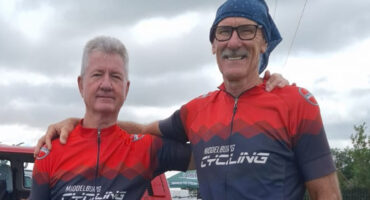 Two elderly men in matching red and blue cycling jerseys stand smiling beside a bike on a grassy field. A red van is parked in the background. Cloudy sky.