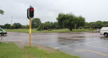 A wet intersection with red traffic lights, indicates that the traffic lights are not working. The intersection is surrounded by lush green grass and trees under a cloudy sky. A van and a distant ambulance can be seen.