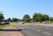 A sunny street scene with traffic lights (not working), a few trees, and cars at an intersection. Clear blue sky and greenery create a calm, open atmosphere.