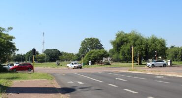 A sunny street scene with traffic lights (not working), a few trees, and cars at an intersection. Clear blue sky and greenery create a calm, open atmosphere.