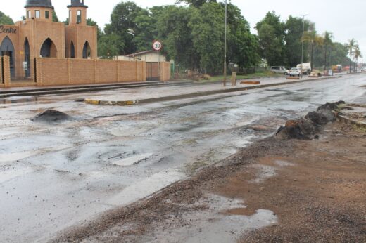 Wet, potholed road with puddles, bordered by a brick building with arched windows. A blurry speed limit sign reads 30. Overcast sky; trees in the background.