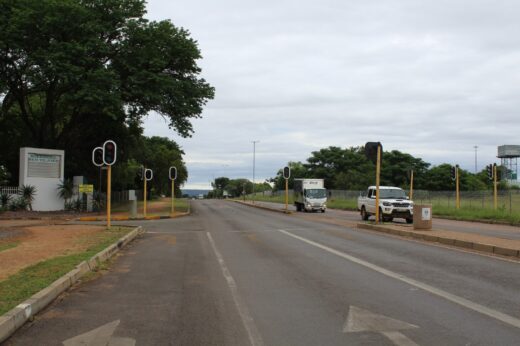 A quiet road flanked by trees and grassy areas, featuring several traffic lights. A white truck and SUV are driving on the right side, under a cloudy sky.