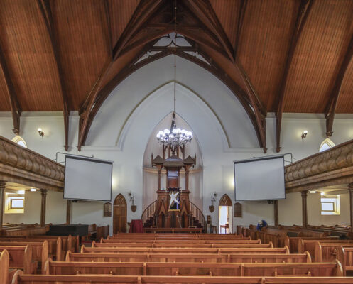 Spacious church interior with wooden pews, arched ceiling, and pulpit. Two large projection screens flank the altar, creating a solemn, peaceful atmosphere.