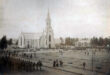 Old photograph of a large church with a tall spire, surrounded by many soldiers in uniform on a parade ground. Civilians gather nearby, creating a historical and solemn atmosphere.