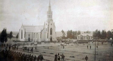 Old photograph of a large church with a tall spire, surrounded by many soldiers in uniform on a parade ground. Civilians gather nearby, creating a historical and solemn atmosphere.