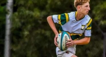 A young rugby player in a gray and green uniform runs on a grassy field, focused intently as he clutches the ball, conveying determination.