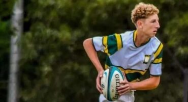 A rugby player in a green and yellow jersey holds a ball, poised to pass. He stands on a grassy field with trees in the background, focused and determined.