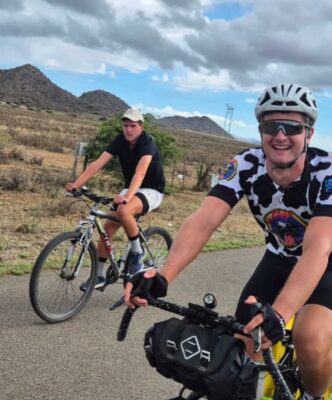 Two cyclists ride on a rural road. The foreground cyclist smiles, wearing a helmet and sunglasses. The background has dry landscape and cloudy skies.