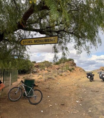 A "STOEL MONUMENT" sign hangs from a tree. Below, a bicycle leans against a trash can. Two motorcycles are parked on a dirt road under a cloudy sky.