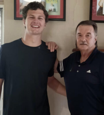 Two men are smiling indoors. The younger man on the left, in a black shirt, has his arm around the older man on the right, wearing a dark polo. Framed photos are on the wall behind them.