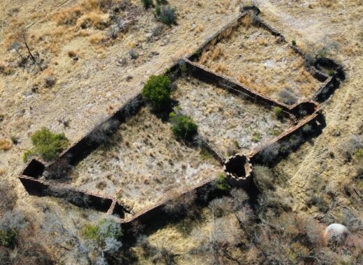 Aerial view of an ancient stone ruin on dry grassland. The walls form a rectangular shape with a round turret, surrounded by sparse vegetation. Rugged, historical feel.