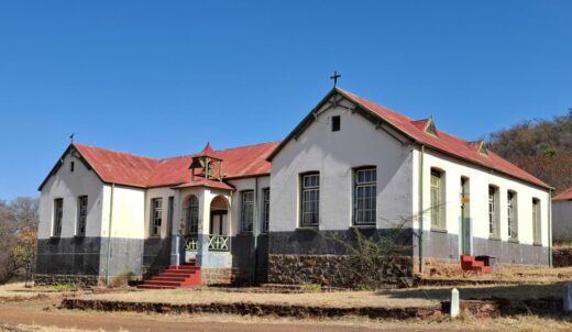 An old house with a red roof and white walls stands under a clear blue sky. It features multiple windows and two crosses, set on a grassy, dry landscape.