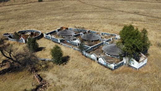 Aerial view of a traditional African homestead featuring round huts with thatched roofs, surrounded by a fence, set in a dry, grassy landscape.