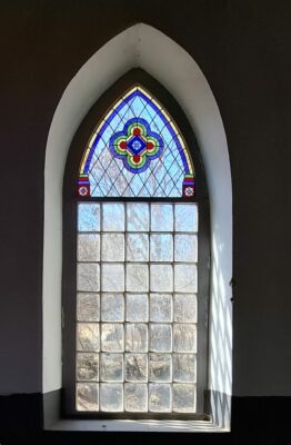 A tall, arched window with colorful stained glass featuring a central geometric pattern. Sunlight filters through clear panes, revealing bare trees outside.