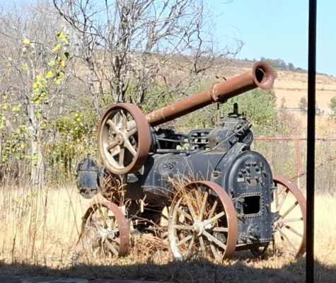 An old, rusted, steam-powered farm machine with large wheels and a long chimney sits overgrown in a grassy field, surrounded by dry, leafless trees.