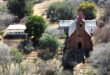 Aerial view of a small, rustic church with a red roof and steeple, surrounded by dry trees and grass. Nearby, there's a modest building with a red roof, giving a serene, rural atmosphere.
