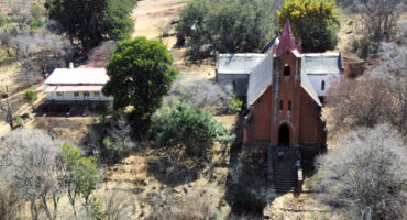 Aerial view of a small, rustic church with a red roof and steeple, surrounded by dry trees and grass. Nearby, there's a modest building with a red roof, giving a serene, rural atmosphere.