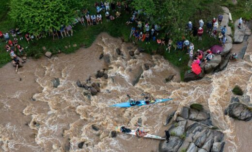 Overhead view of kayakers navigating a turbulent, rocky river. A large group of spectators watch from the grassy riverbank. The scene is dynamic and lively.